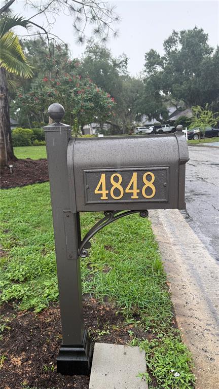 A dark gray metal mailbox with gold house numbers stands on a post near a road — 4848 HAWKSHEAD PARK, SARASOTA, FL
