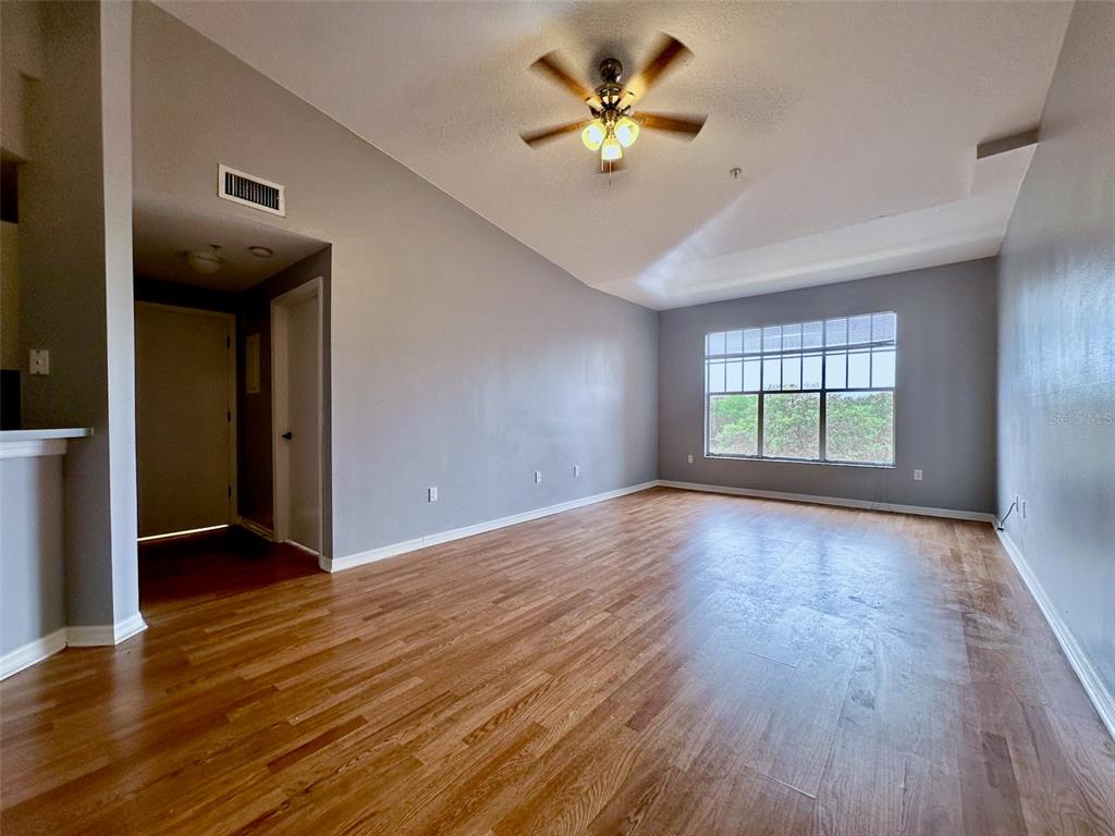 An empty living room features hardwood floors, gray walls, a ceiling fan, and a large window — 5578 METROWEST BOULEVARD UNIT 1-302, ORLANDO, FL