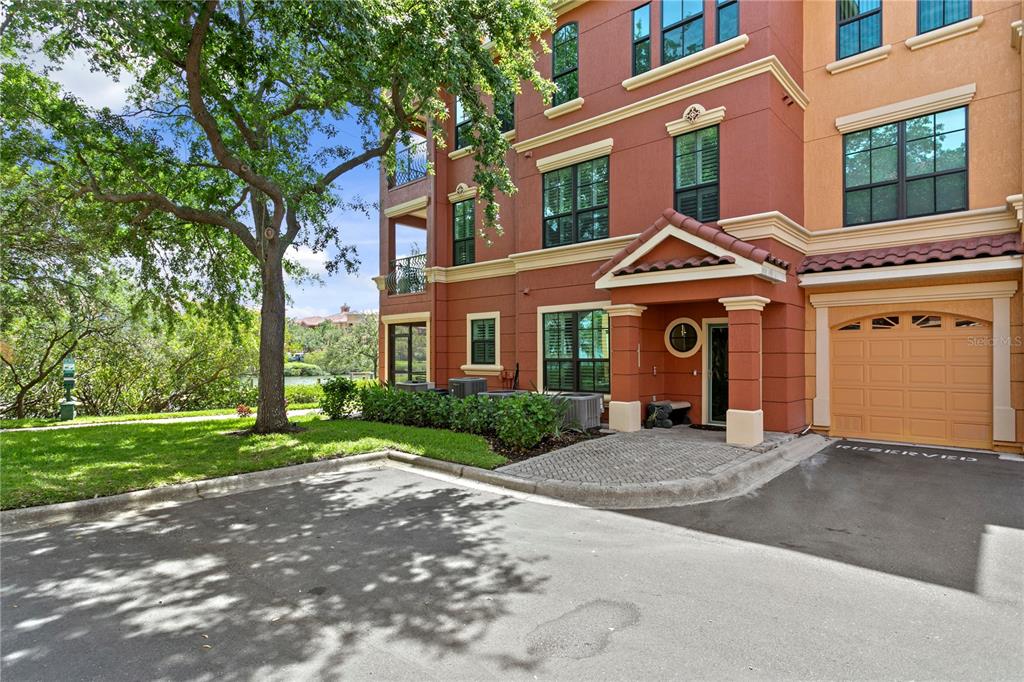 A three-story townhouse with a tan garage, red stucco exterior, and a paved entryway — 2741 VIA CIPRIANI UNIT 914A, CLEARWATER, FL