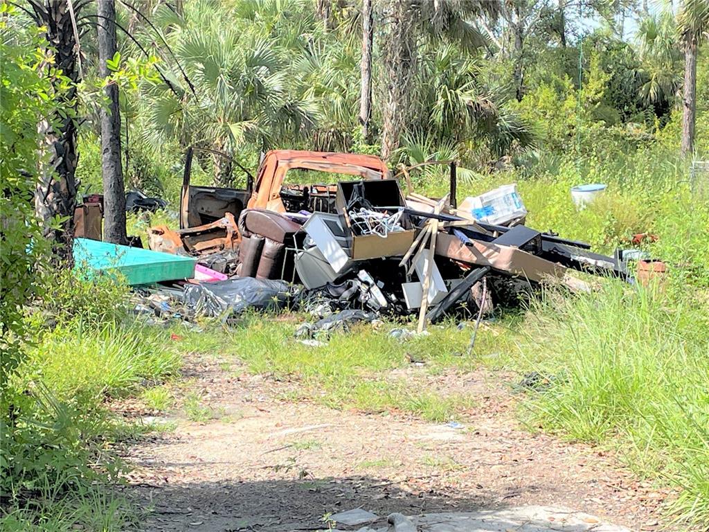 A pile of trash and a burnt-out vehicle frame sit in a grassy, wooded area — 1823 INSPIRATION COURT, LEHIGH ACRES, FL