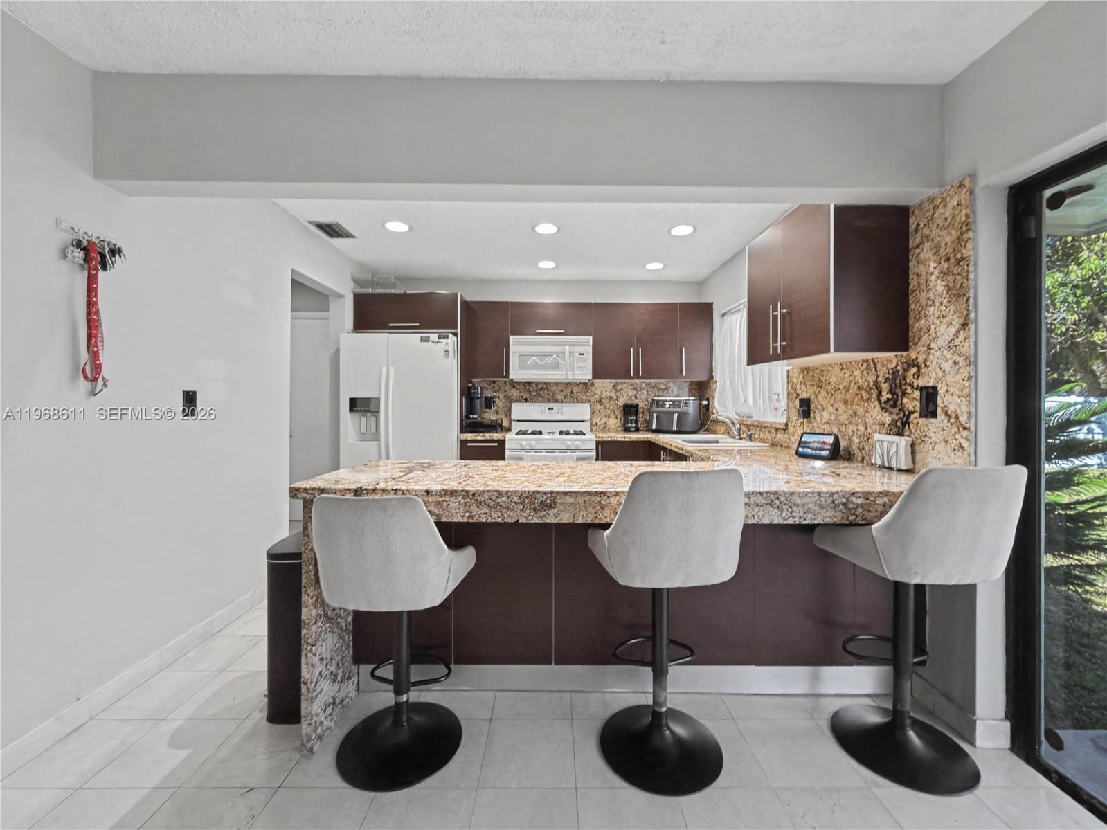 A kitchen features dark wood cabinets, granite countertops, a breakfast bar, and three bar stools — 10491 SW 122nd Ct, Miami FL 33186, MIAMI, FL