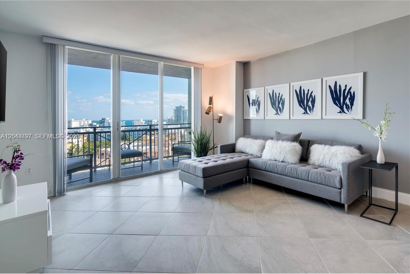 A modern living room features a gray sectional sofa, tiled floors, and a balcony with ocean views — 90 Alton Rd # 1207, Miami Beach FL 33139, MIAMI BEACH, FL