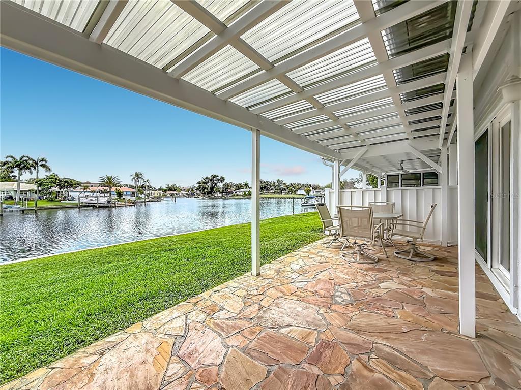 A stone-paved patio with a covered roof overlooks a grassy lawn and a canal — 6537 CAPE HATTERAS WAY NE UNIT 2, ST PETERSBURG, FL