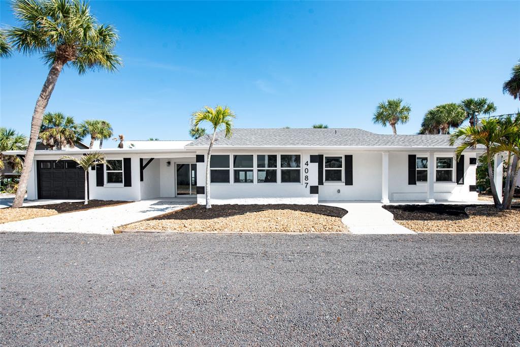 A single-story white house with black shutters and a gravel driveway features palm trees — 4087 PELICAN SHORES CIRCLE, ENGLEWOOD, FL
