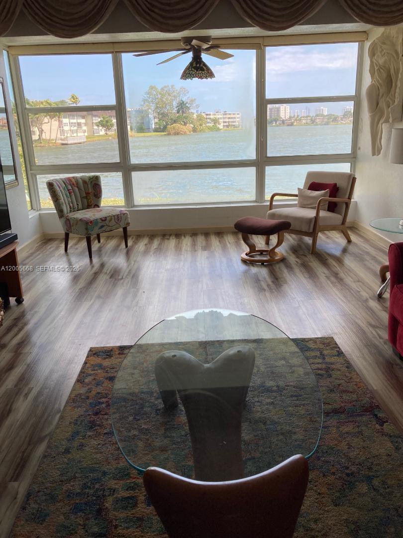 A living room features wood-look flooring, a glass coffee table, and large windows overlooking water — 18901 NE 14th Ave # 101, Miami FL 33179, MIAMI, FL