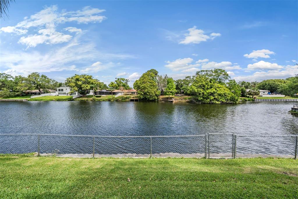 A grassy backyard with a chain-link fence overlooks a calm lake and distant houses — 2424 FAIRBANKS DRIVE, CLEARWATER, FL