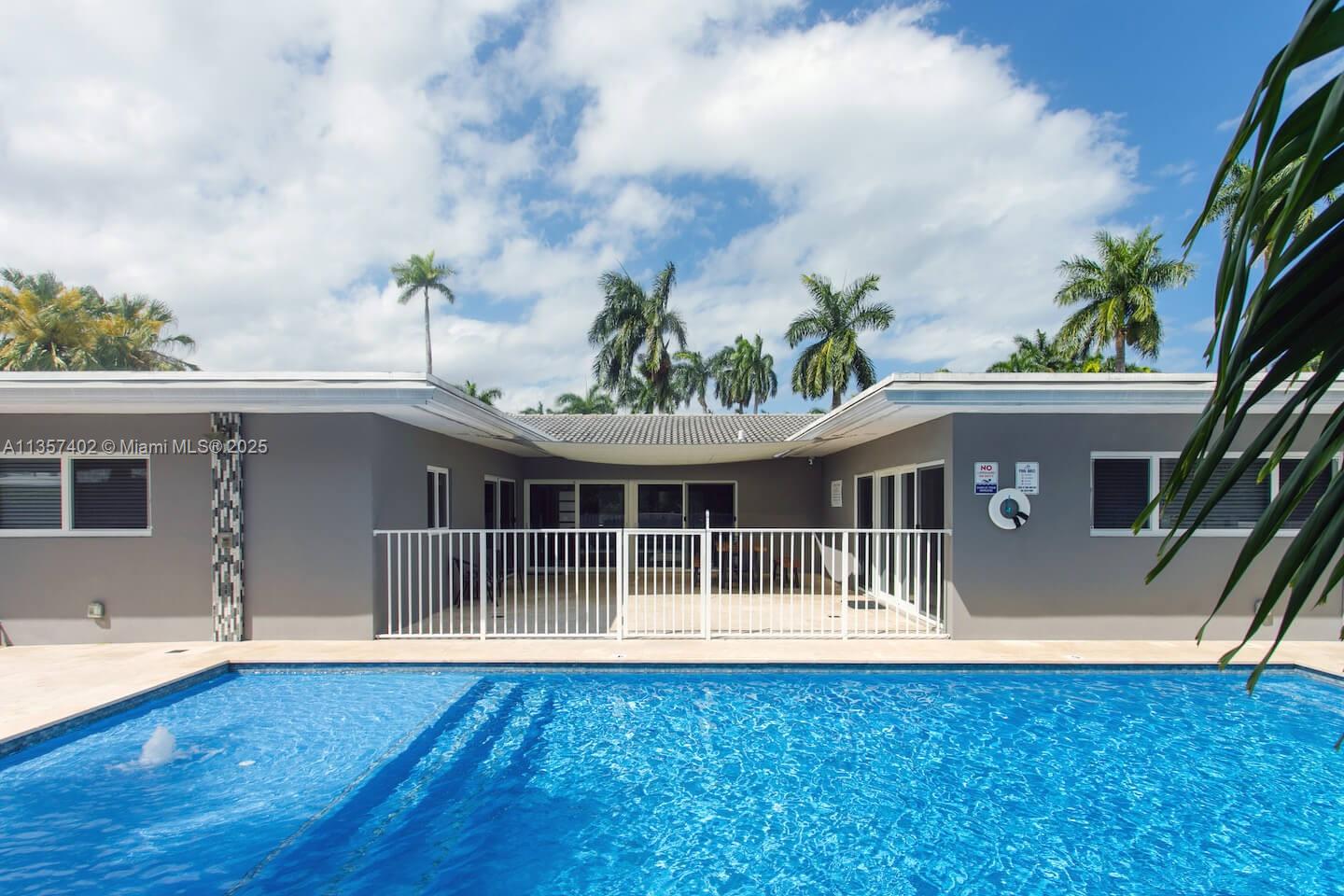 A swimming pool sits in front of a gray house with a white fence and patio — 1218 Hollywood Blvd, Hollywood FL 33019, HOLLYWOOD, FL